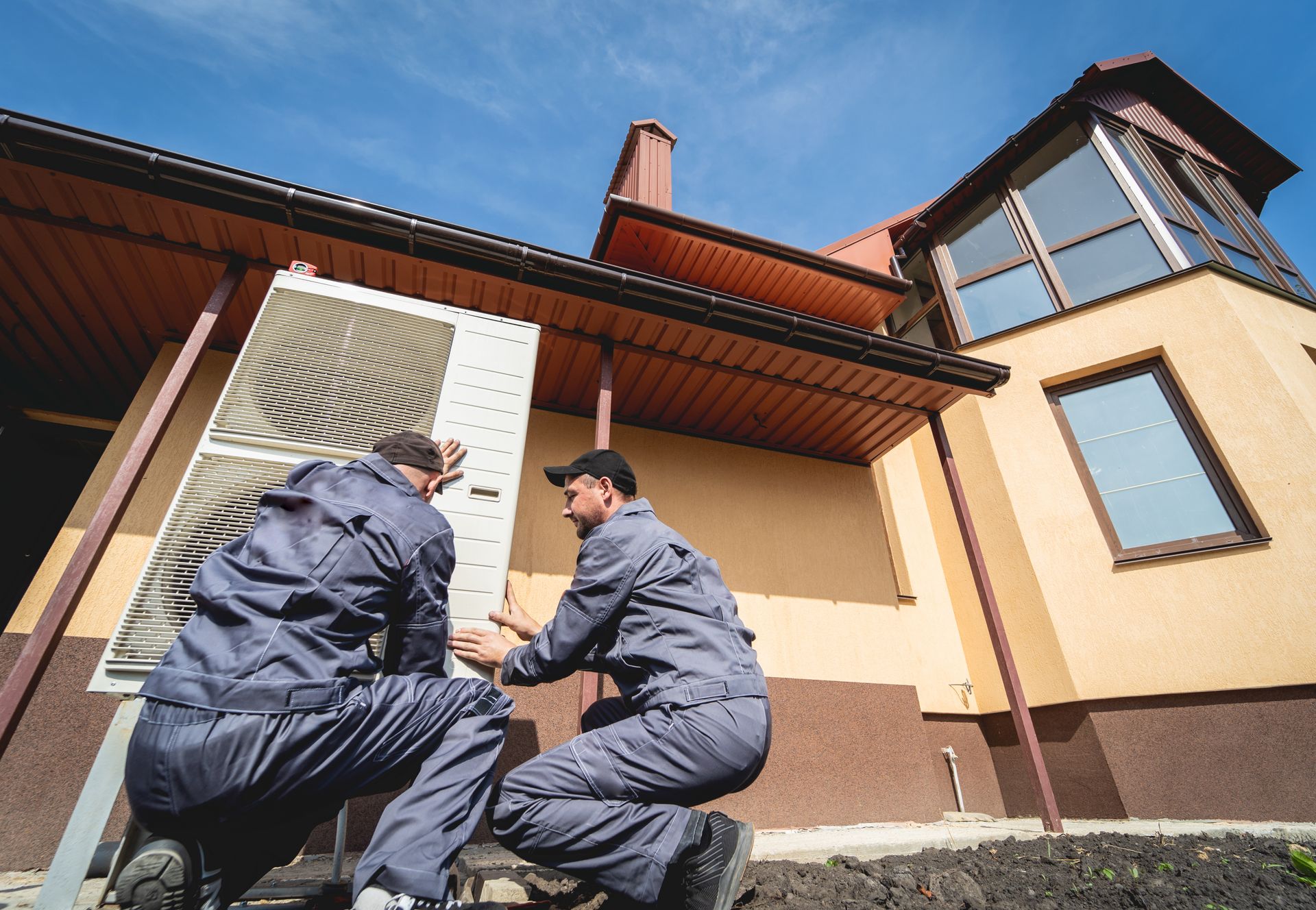 Two workers in gray uniforms kneeling outside a house while installing an outdoor air conditioning unit.