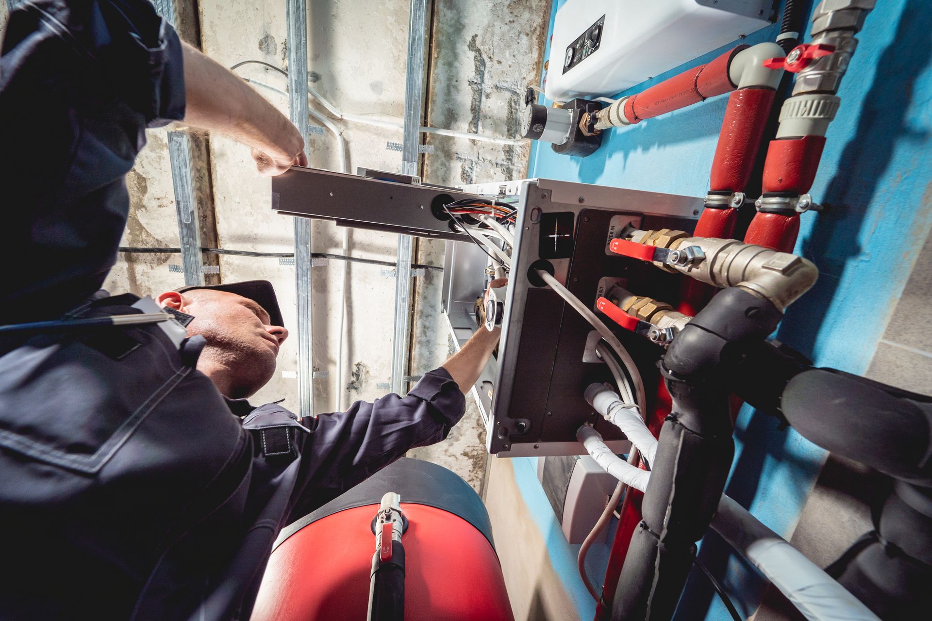 Plumber working on pipes and electrical wiring near a blue wall.