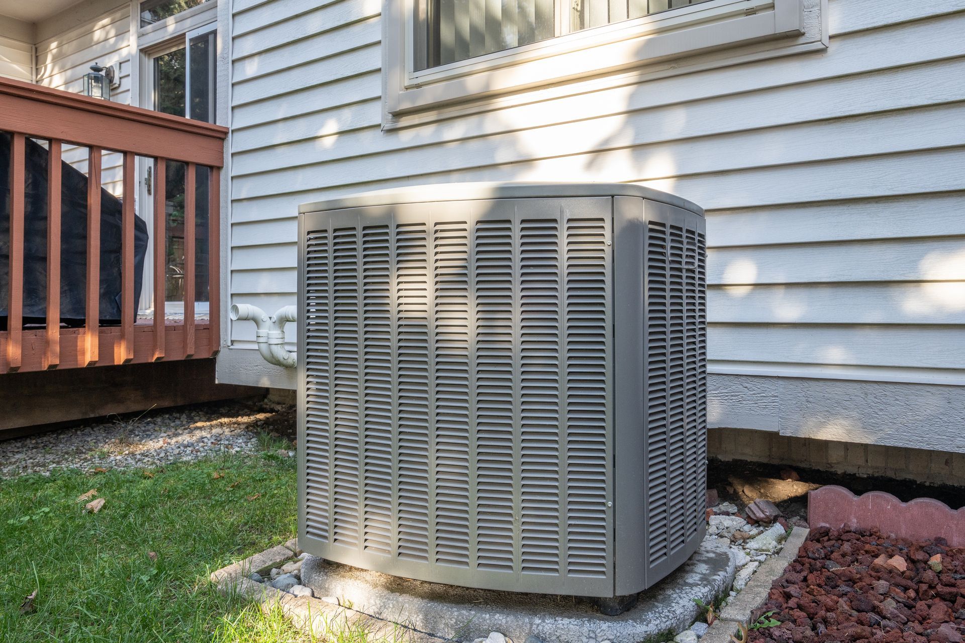 Outdoor air conditioning unit next to a house with a wooden deck and green grass.