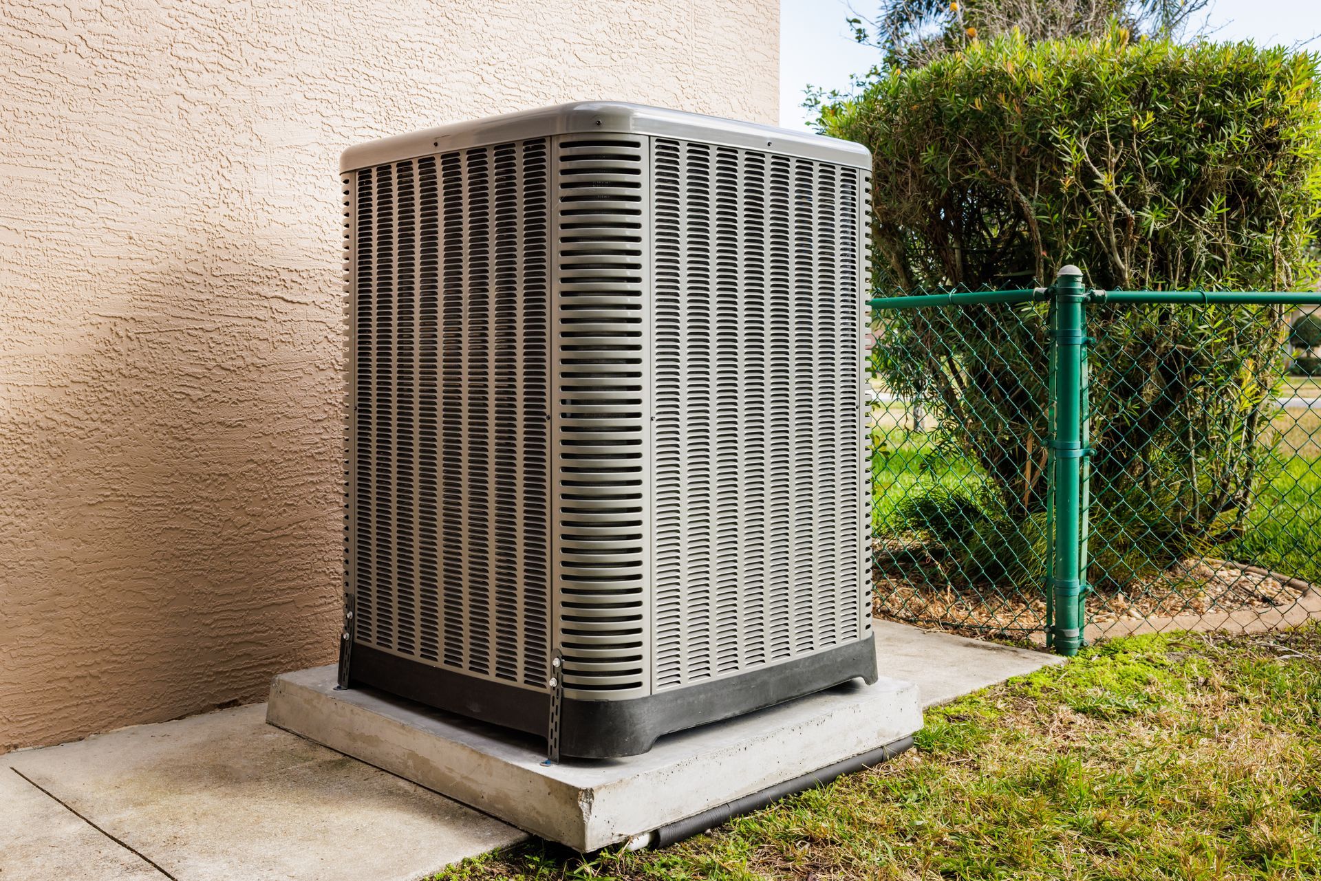 A square, grey outdoor air conditioning unit sits on a concrete pad next to a beige wall and a green chain-link fence.