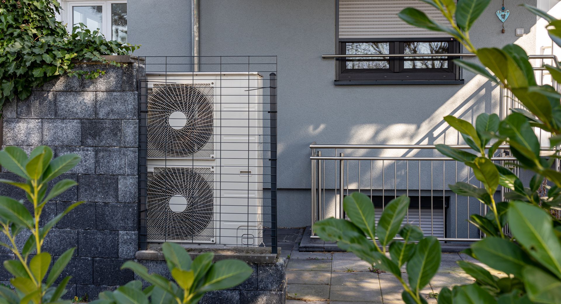A white dual-fan heat pump unit sits inside a black metal cage beside a grey house wall with a basement window. A white dual-fan heat pump unit sits inside a black metal cage beside a grey house wall with a basement window.