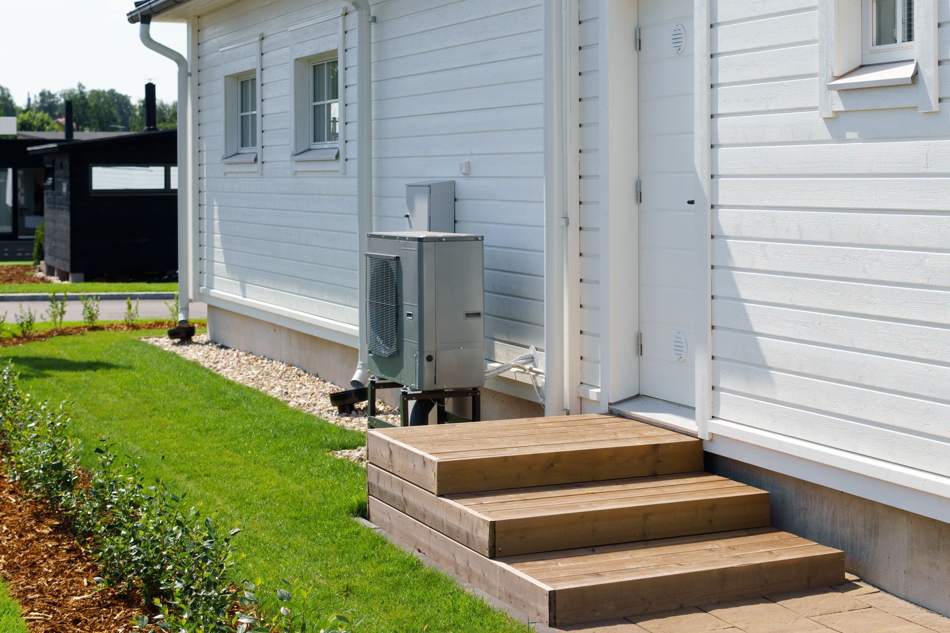 A gray heat pump unit stands beside the wooden steps leading to a white, horizontal-sided house with a manicured lawn. A gray heat pump unit stands beside the wooden steps leading to a white, horizontal-sided house with a manicured lawn.