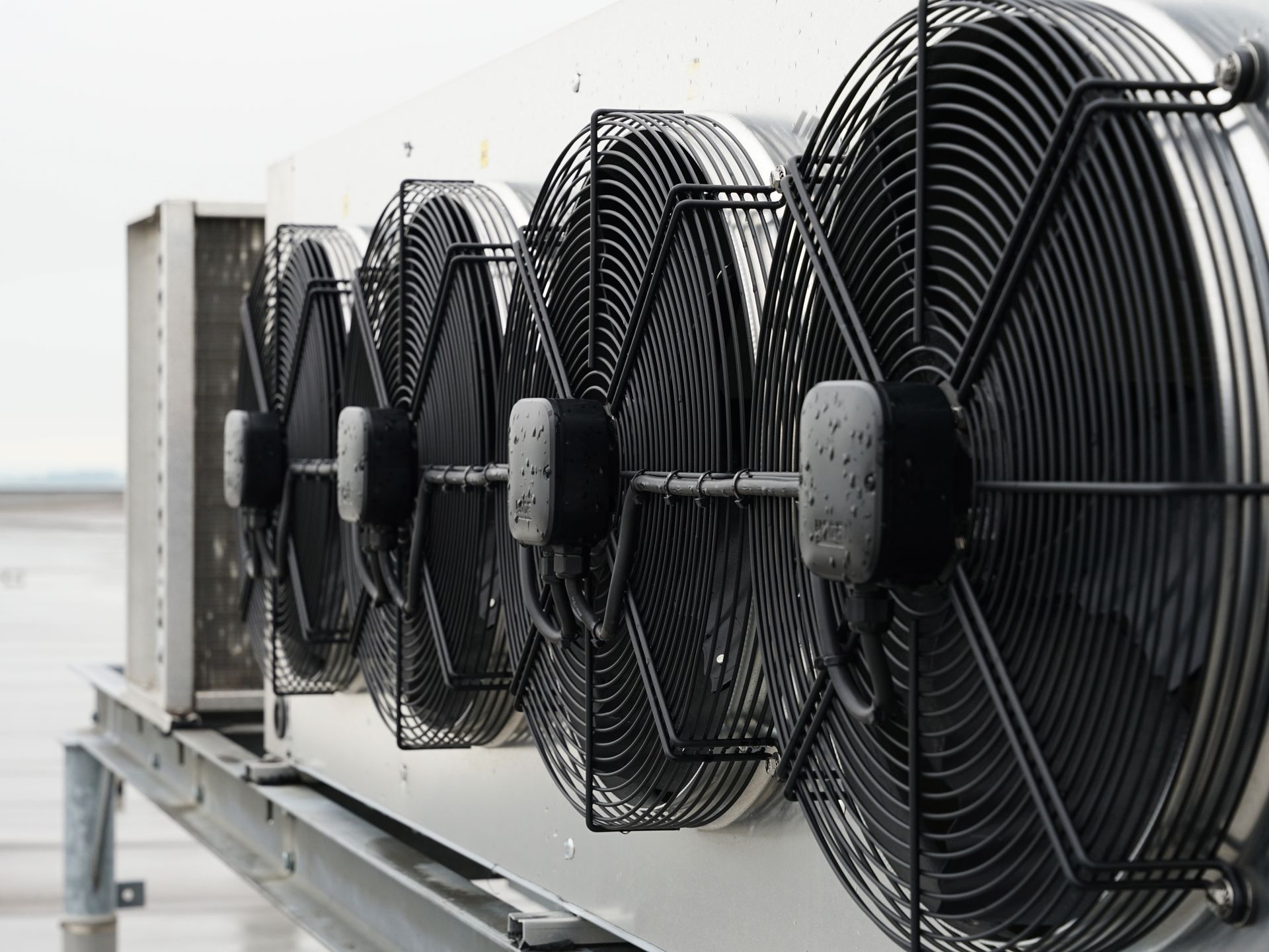 A row of industrial cooling fans with black wire grilles mounted on a metal unit on a rooftop. A row of industrial cooling fans with black wire grilles mounted on a metal unit on a rooftop.