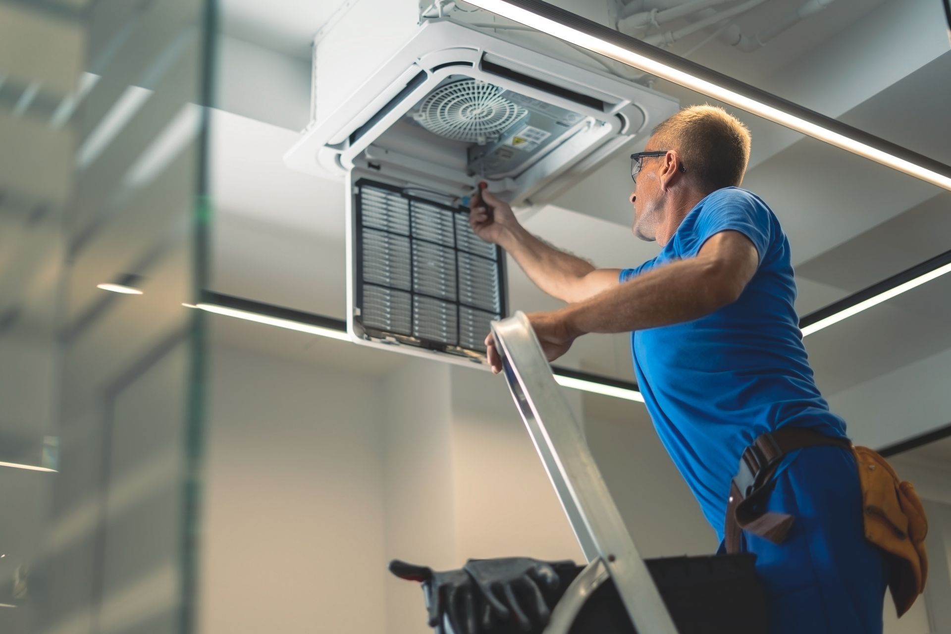 Man on a ladder cleaning an air conditioning filter in a modern office.