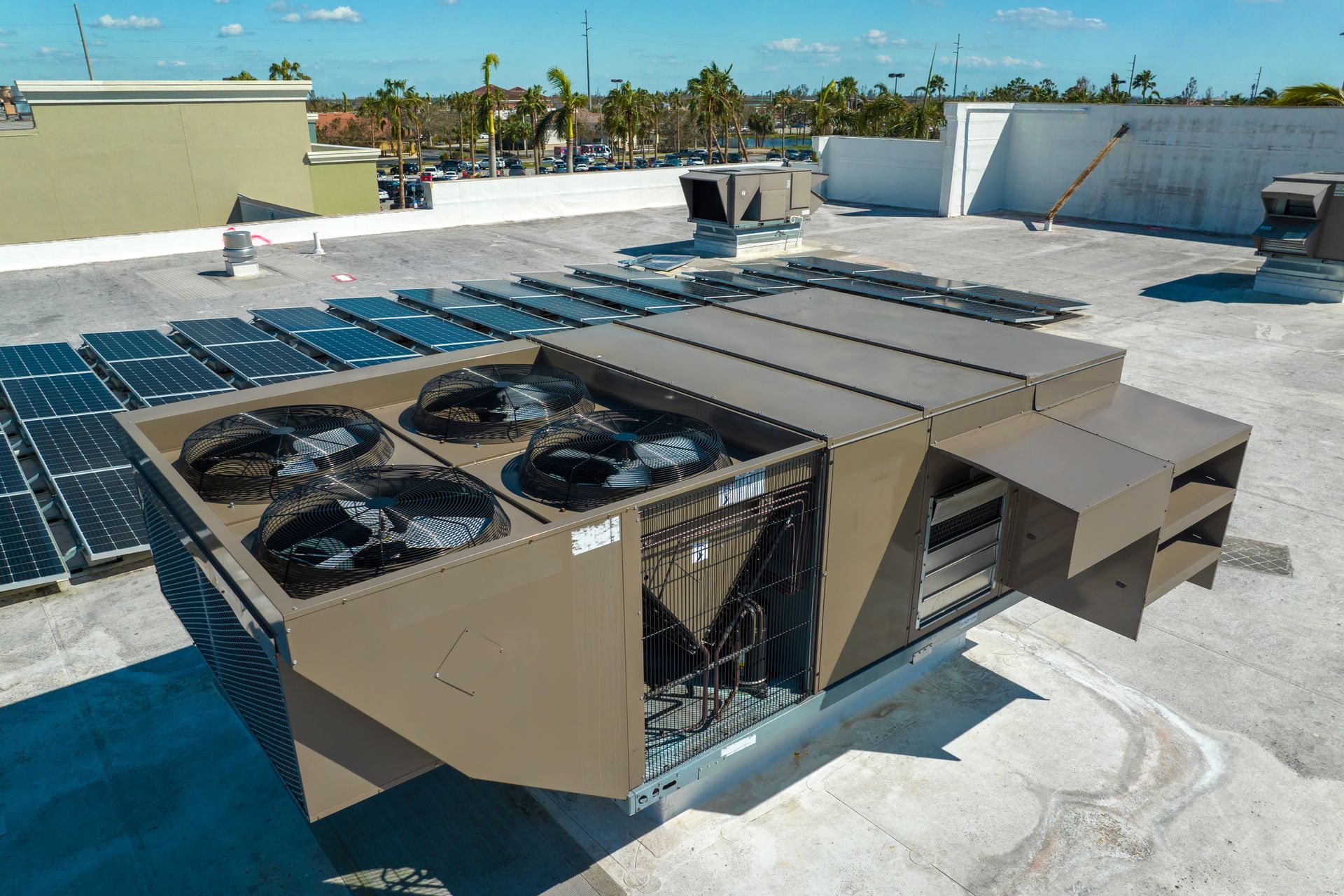 A beige rooftop HVAC unit with four fans sits on a flat building roof next to rows of solar panels under a clear blue sky. A beige rooftop HVAC unit with four fans sits on a flat building roof next to rows of solar panels under a clear blue sky.