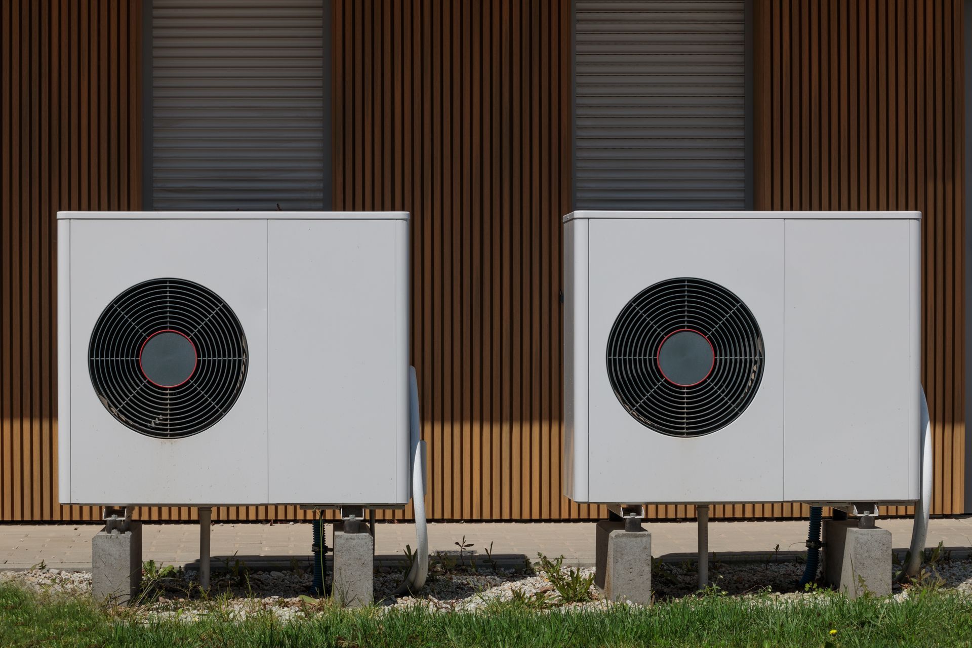 Two white heat pump units sit on concrete blocks against a brown, vertically slatted building wall.