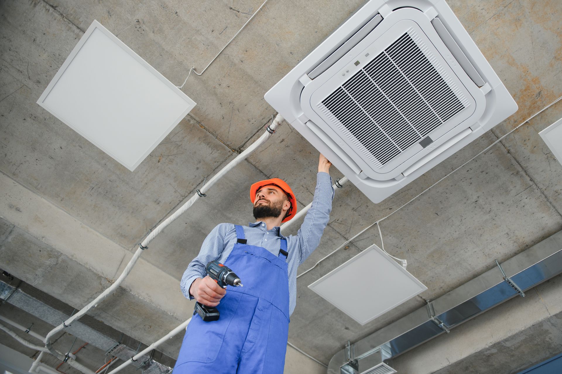 A worker in a hard hat and blue coveralls uses a drill to install a ceiling-mounted air conditioning unit. A worker in a hard hat and blue coveralls uses a drill to install a ceiling-mounted air conditioning unit.