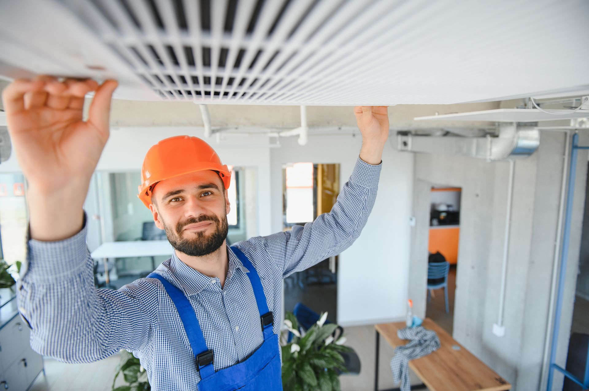 A person in an orange hard hat and blue coveralls installing a ventilation grate on a ceiling in a modern office space. A person in an orange hard hat and blue coveralls installing a ventilation grate on a ceiling in a modern office space.