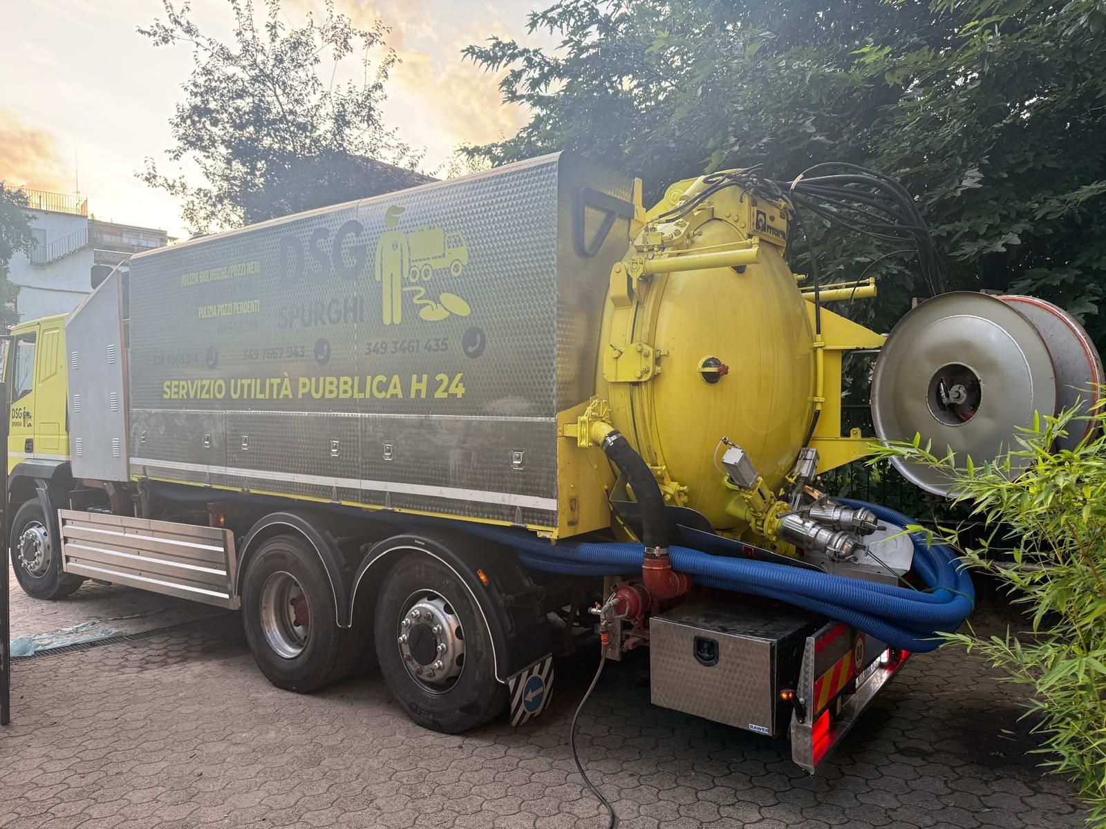 Un camion della nettezza urbana giallo e grigio parcheggiato all'aperto, con tubi blu e alberi sullo sfondo.