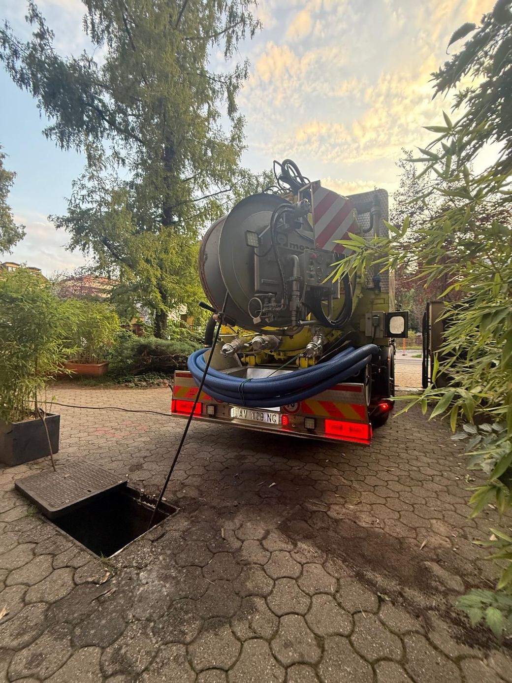 Un camion fognario che serve un tombino aperto su un'area pavimentata in mattoni, sotto un cielo nuvoloso.