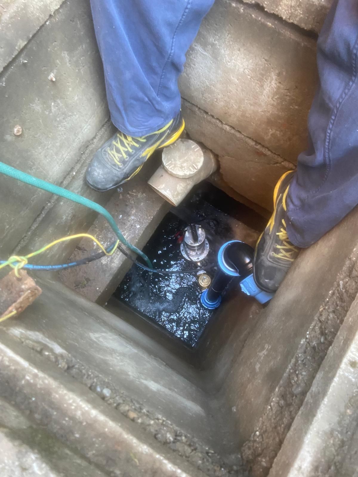 Persona che lavora all'interno di un pozzo di cemento. Acqua scura e tubi all'interno. La persona indossa pantaloni blu e scarpe gialle e nere.