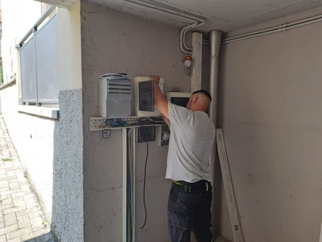 Un uomo installa componenti elettrici su un muro di cemento all'aperto. Sono presenti elementi grigi, neri e metallici.