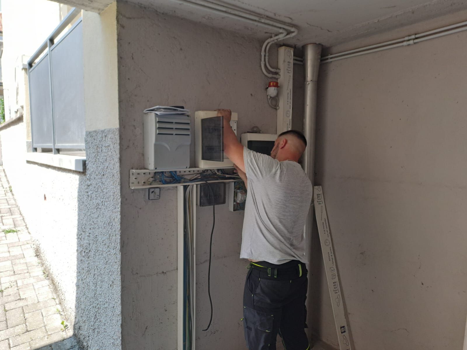Un uomo installa componenti elettrici su un muro di cemento all'aperto. Sono presenti elementi grigi, neri e metallici.
