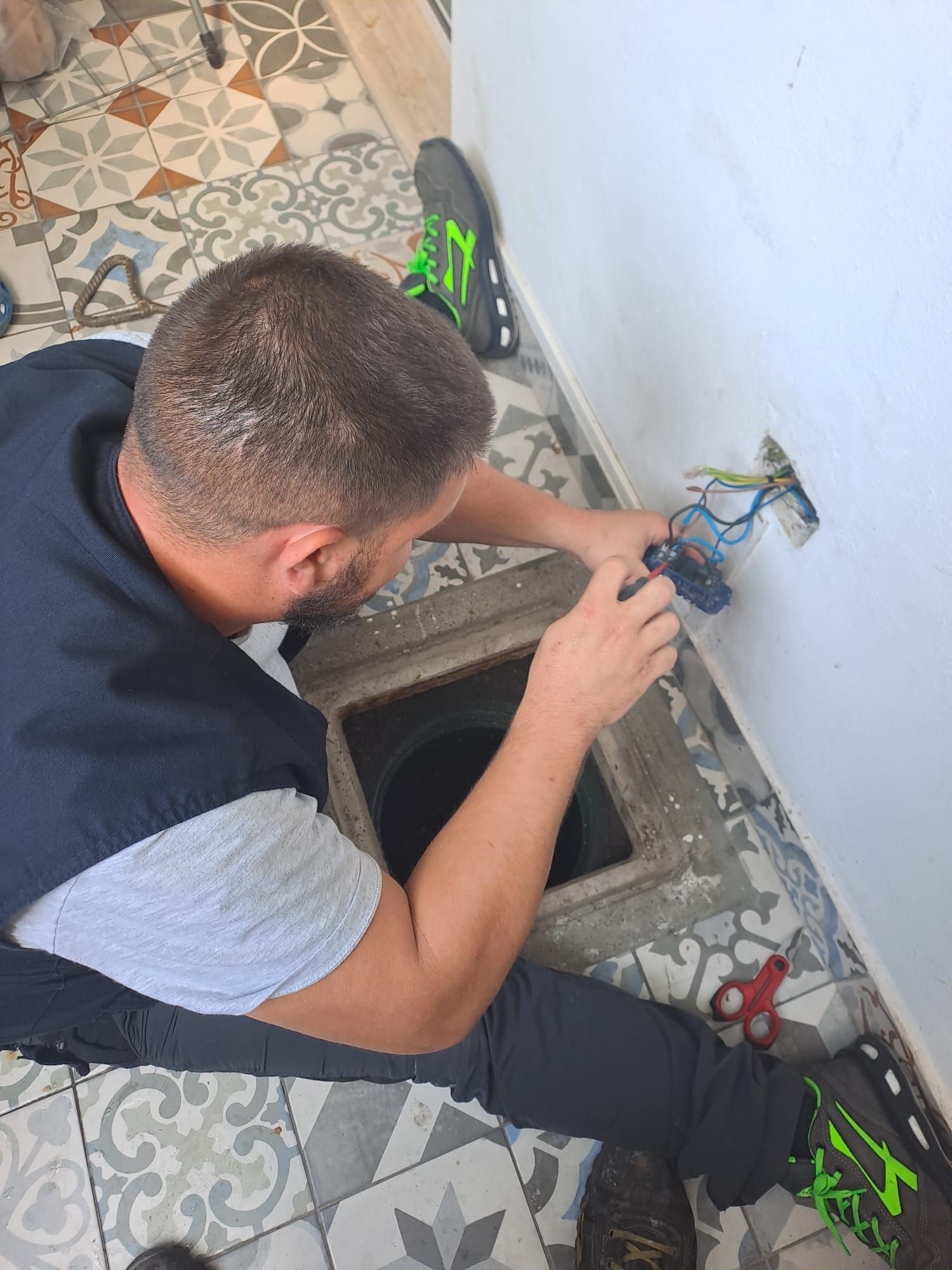 Uomo che lavora su un impianto elettrico, vicino a uno scarico a pavimento, con degli attrezzi sul pavimento piastrellato.