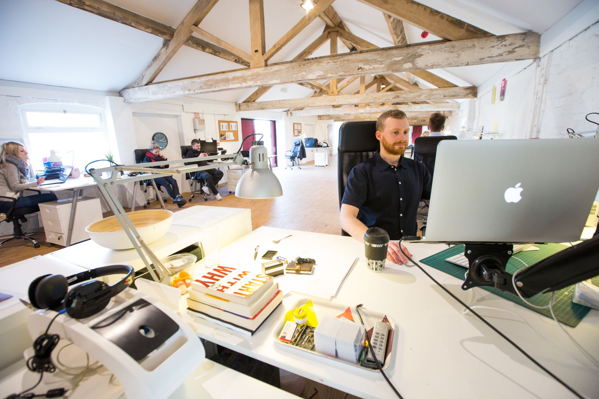 Office with people at desks, working on computers. Man in blue shirt uses laptop. White walls, wood beams.