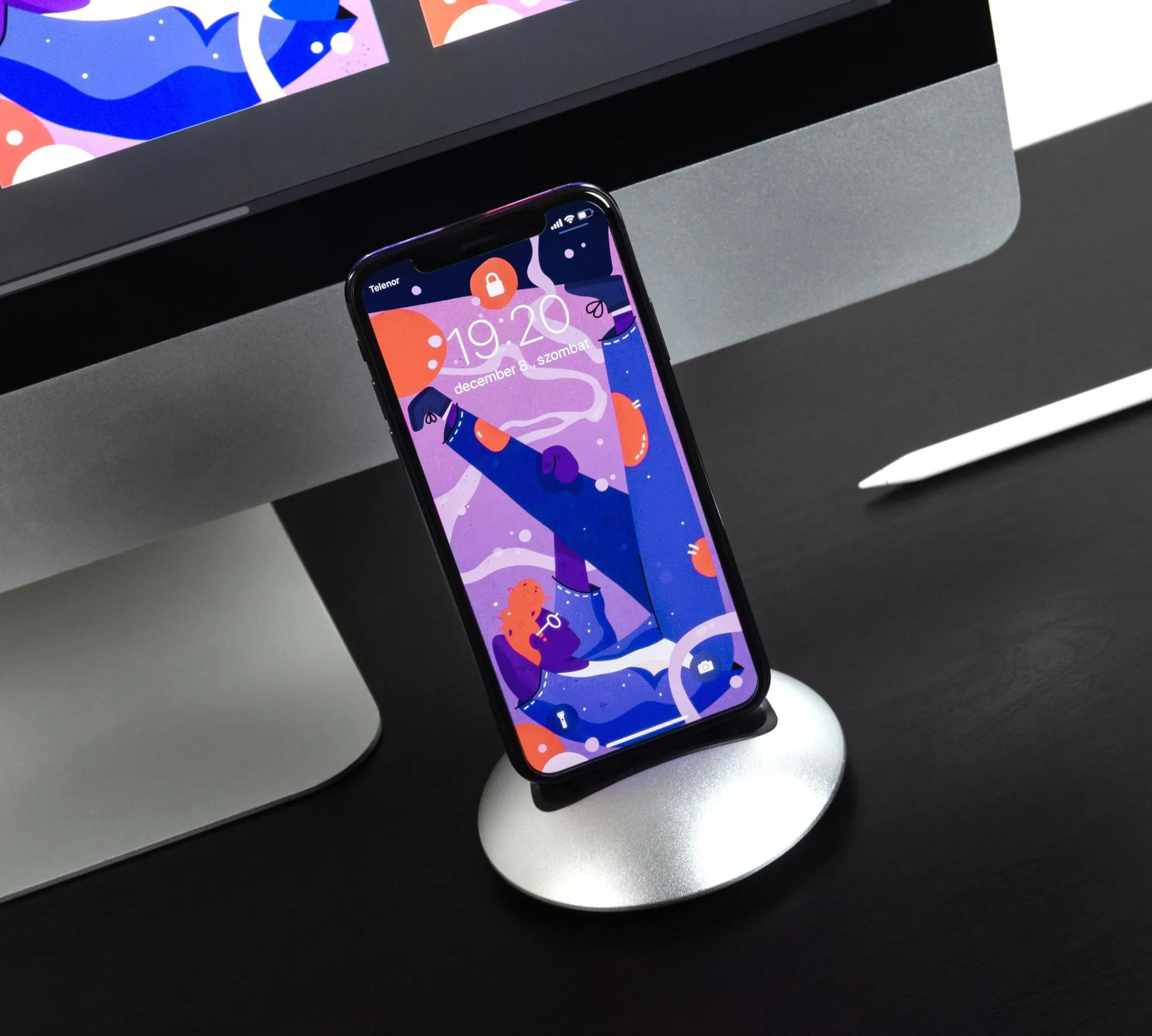 Smartphone on a silver stand displaying a colorful lock screen, next to a computer monitor and a white stylus.