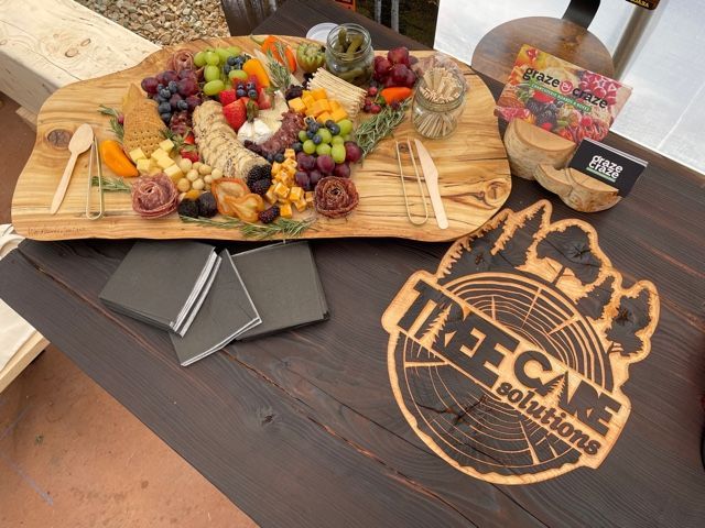 Charcuterie board with fruit, cheese, and meats, next to napkins and a Tree Care Solutions logo on a wooden table.
