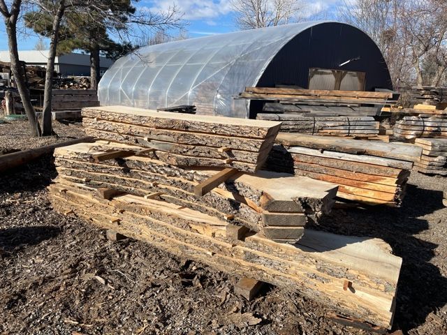 Stacks of rough-hewn lumber sit outdoors near a translucent-roofed structure.