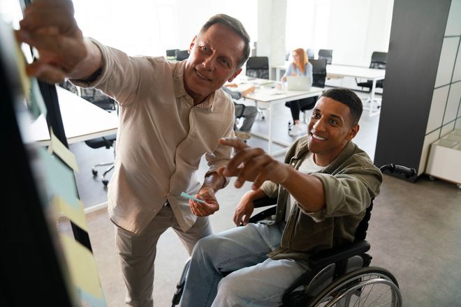Two men in an office reviewing notes on a board; one in a wheelchair points, the other gestures.