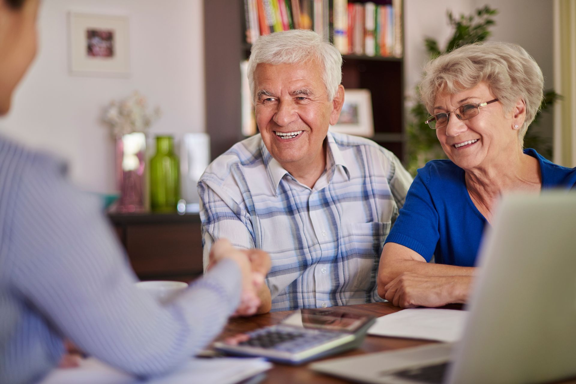 Senior couple smiling and shaking hands with a person at a table, possibly financial advisor, at home.