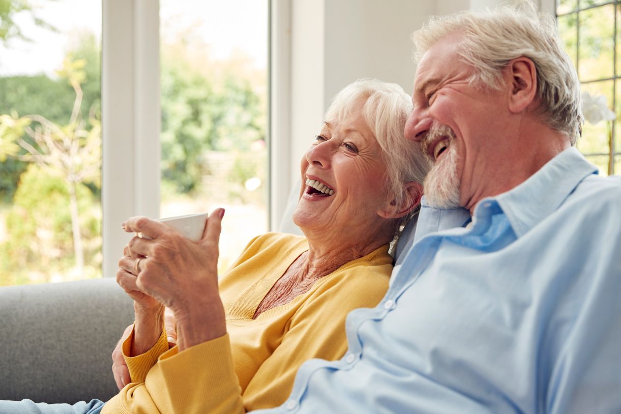 An elderly couple is sitting on a couch laughing and drinking coffee.