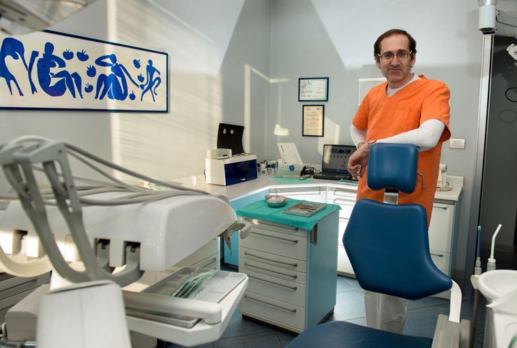 Dentista in camice arancione in piedi in uno studio dentistico. Sono visibili una poltrona blu, attrezzature, opere d'arte e una finestra.