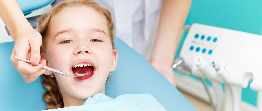 Una ragazzina dal dentista. Il dentista le controlla i denti con uno strumento di metallo. Lei apre la bocca e sorride.