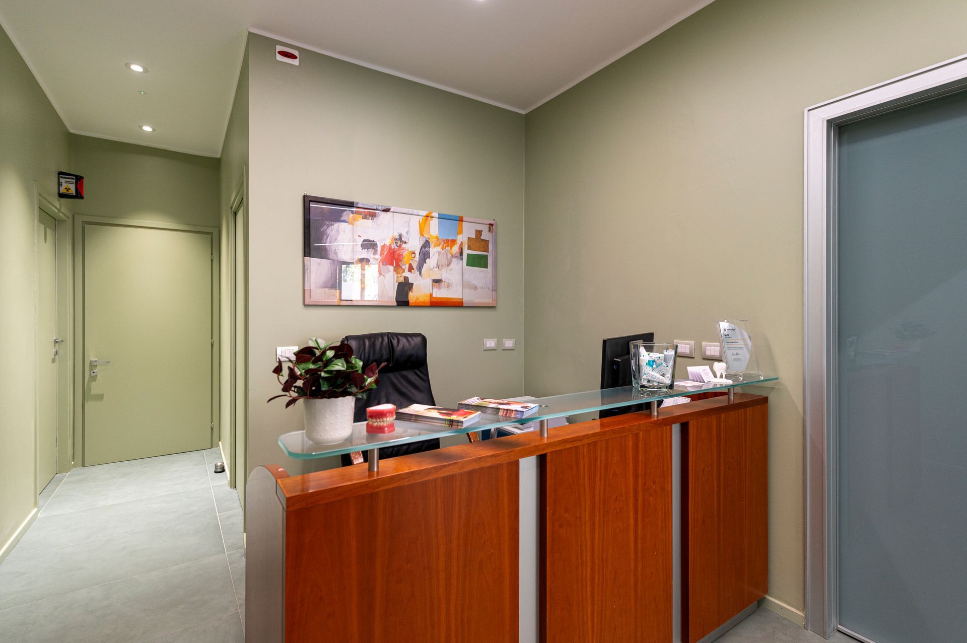 Area reception con scrivania in legno, opere d'arte e porta d'ingresso. Pareti verde oliva, luci soffuse e design moderno.