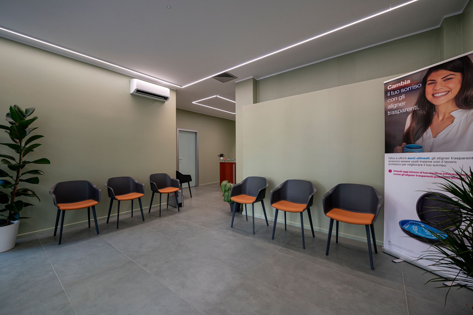 Sala d'attesa con sedie, una pianta e un poster. Pareti verde chiaro, pavimento grigio e illuminazione dall'alto.