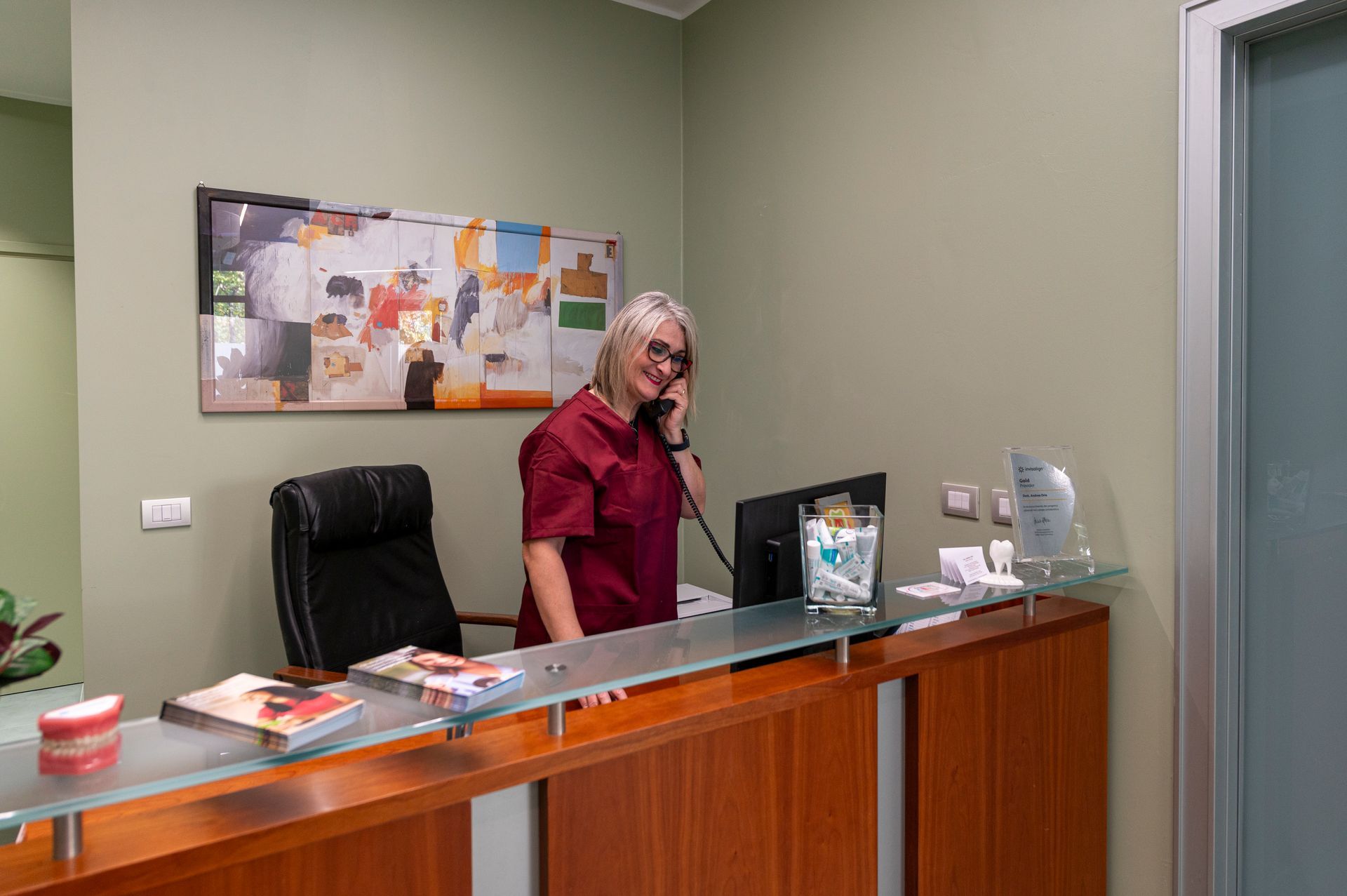 Una receptionist in camice color bordeaux risponde al telefono alla reception di uno studio dentistico.