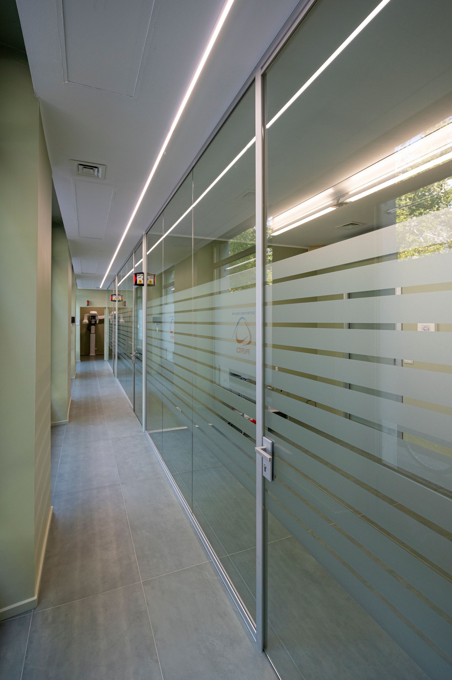 Corridoio con pareti in vetro caratterizzate da strisce orizzontali satinate e un lampadario sul soffitto.