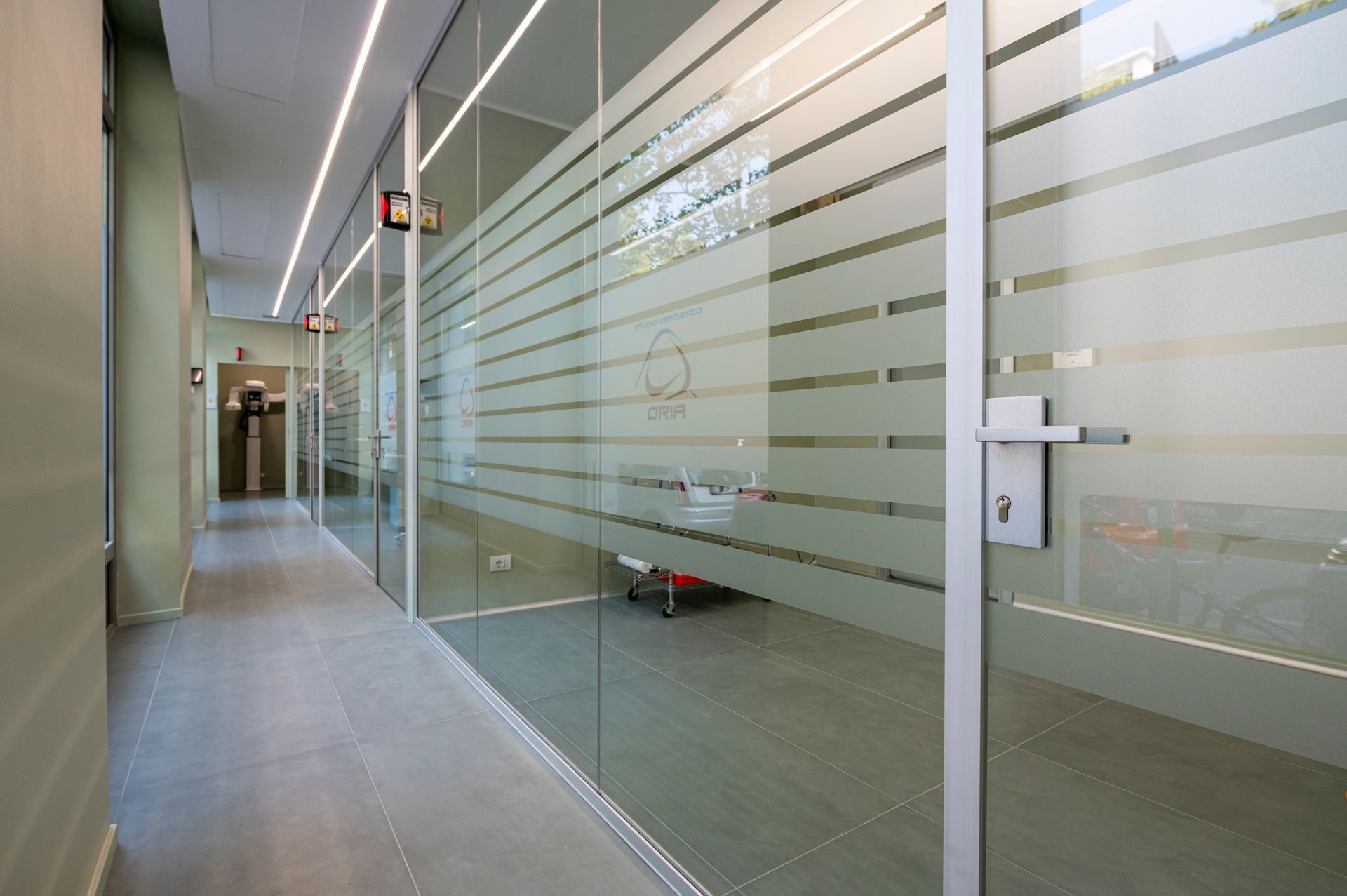 Corridoio con uffici con pareti in vetro. Strisce orizzontali satinate garantiscono la privacy. Illuminazione fluorescente, pavimento grigio e una porta sulla destra.