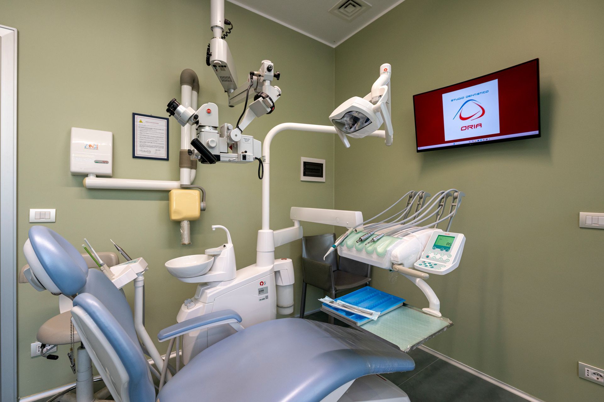 Studio dentistico con poltrona, attrezzatura e monitor montato. Pareti verdi, poltrona grigia.
