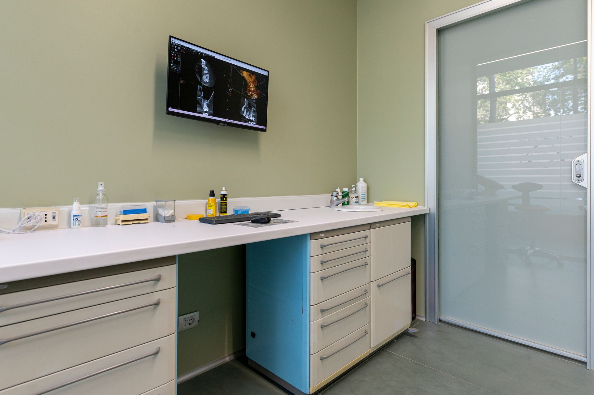 Sala visite con pareti verde chiaro, bancone bianco e armadietti blu. Una TV trasmette un'immagine.