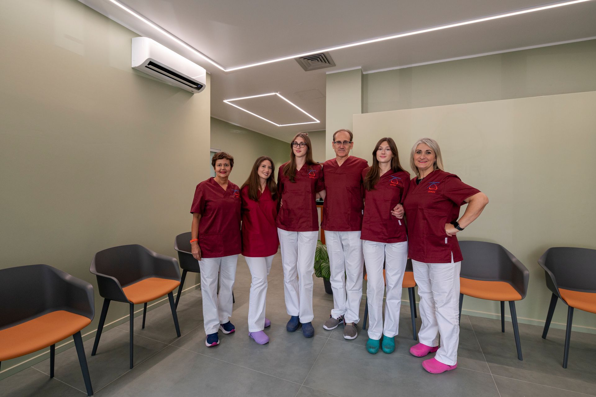 Personale medico in camice bordeaux e pantaloni bianchi posa in una sala d'attesa.