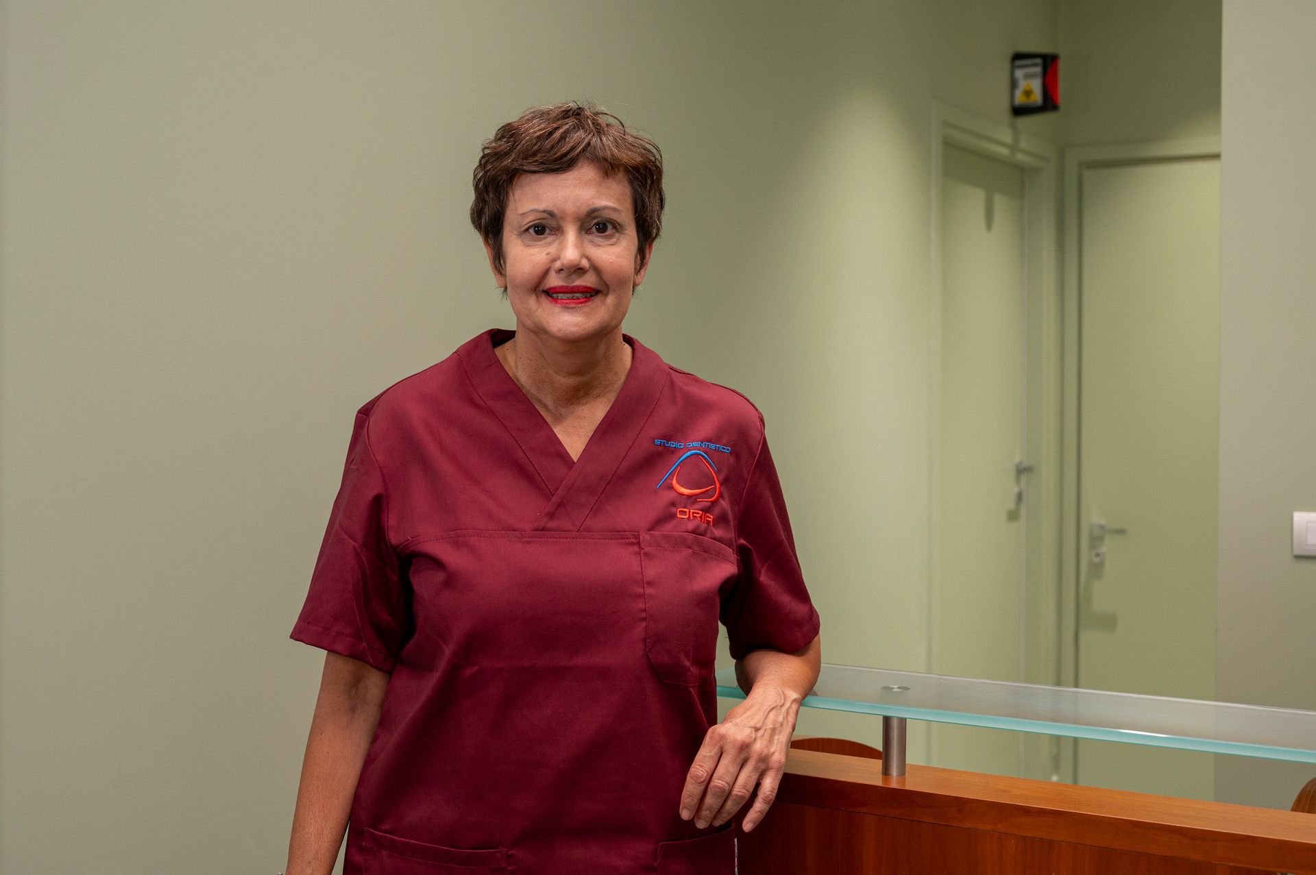 Donna in camice color marrone alla scrivania di uno studio medico, sorridente.