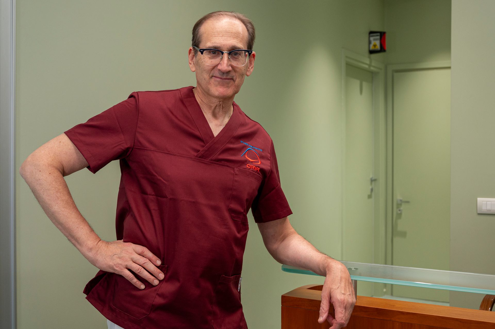 Uomo in camice color bordeaux, occhiali, appoggiato al bancone di uno studio medico, mani sui fianchi.