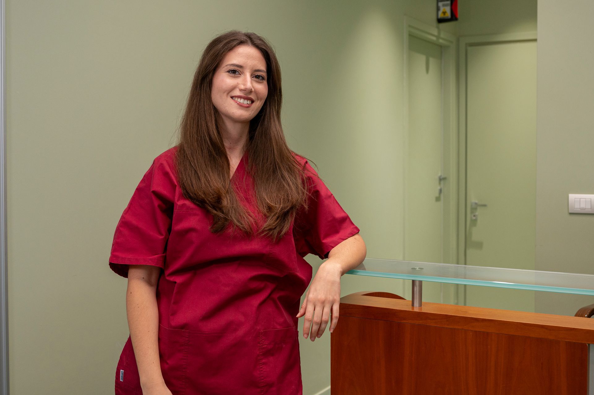 Una donna in camice color bordeaux sorride, appoggiata a un bancone di legno in uno studio medico.