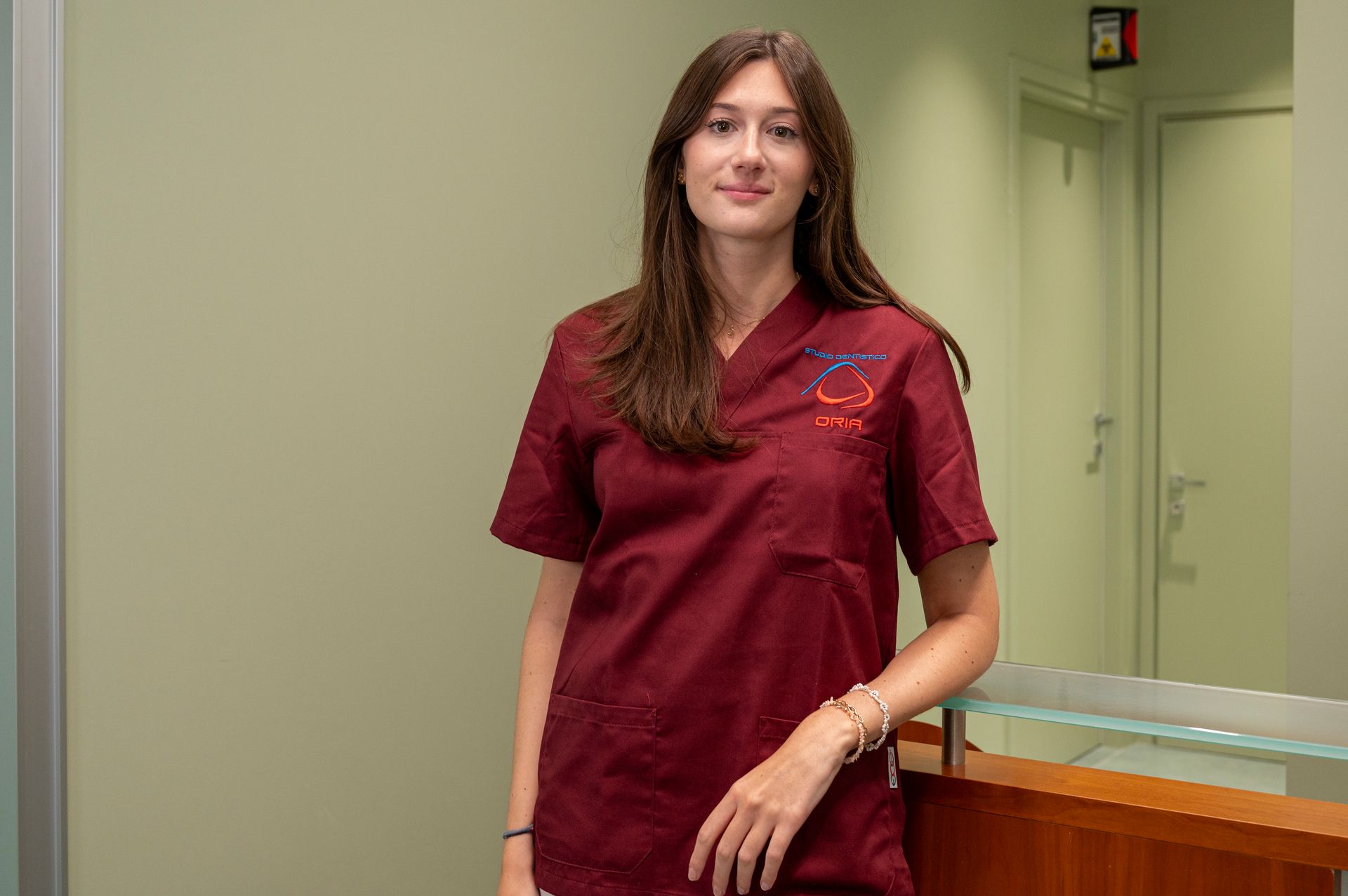 Una donna in camice bordeaux è in piedi accanto al bancone di uno studio medico e sorride.