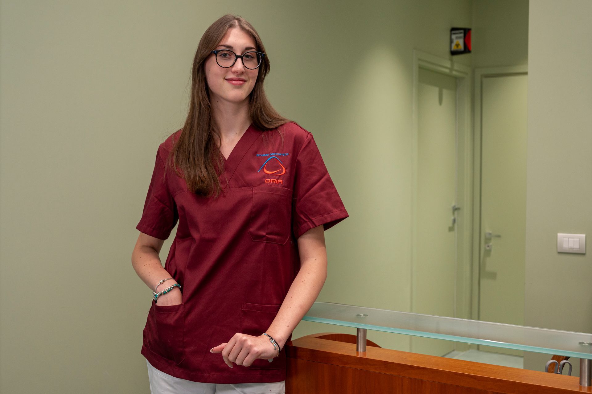 Donna in camice color castagna con occhiali, in piedi alla reception.