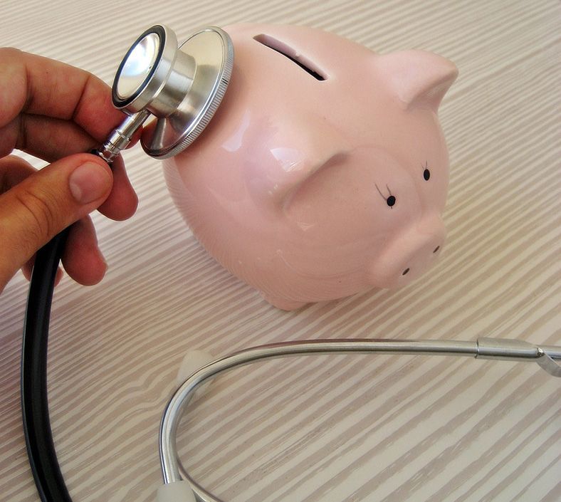 Mano che tiene uno stetoscopio vicino a un salvadanaio rosa, a indicare un controllo della salute finanziaria.