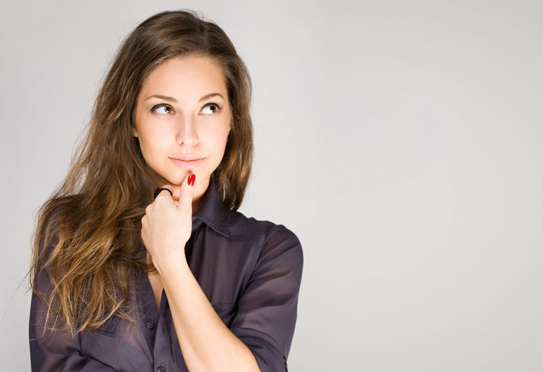 Donna con capelli castani ondulati, sorridente, sguardo laterale, mano sul mento. Su sfondo grigio.