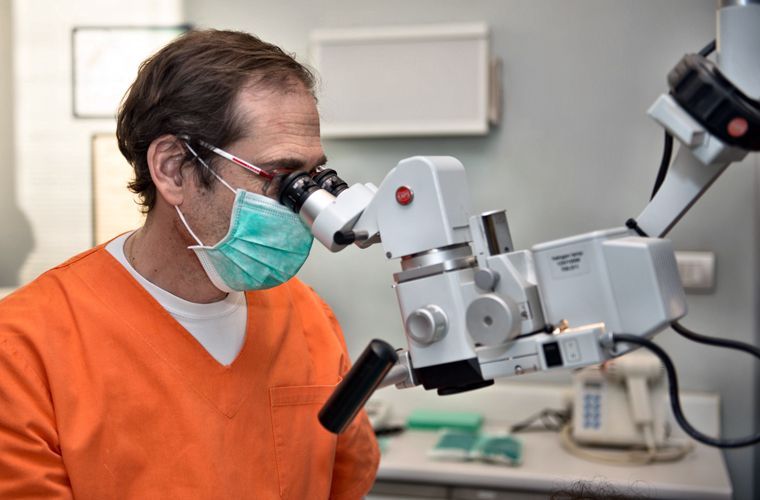 Medico in camice arancione con mascherina, che osserva al microscopio chirurgico. Ambiente di studio medico.