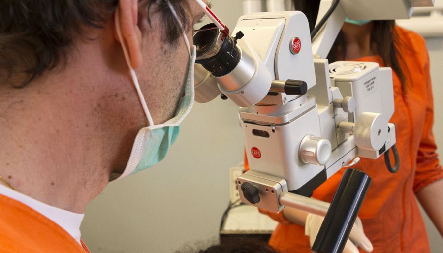 Un dentista che guarda al microscopio, indossando una mascherina. Un'altra persona vestita di arancione è in piedi lì vicino.
