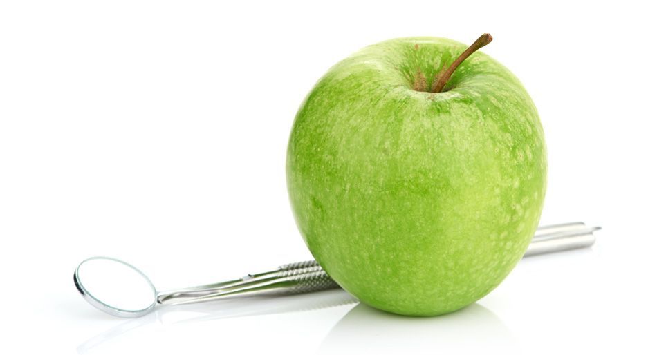 Mela verde accanto a uno specchietto dentale su una superficie bianca.