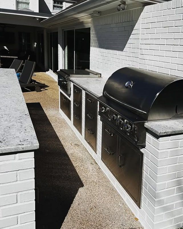 Outdoor kitchen with stainless steel grill and cabinetry against a white brick wall.