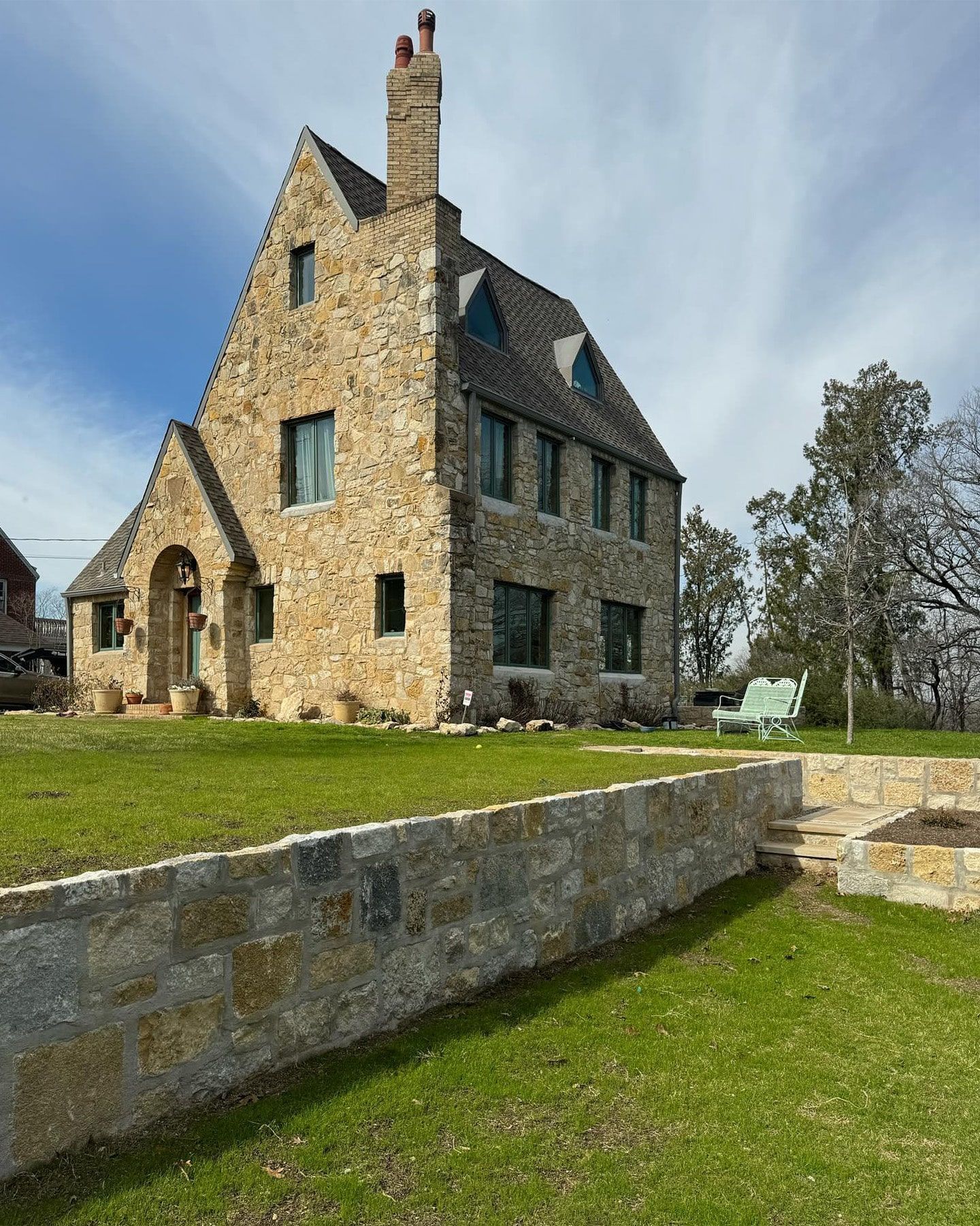 Stone house with steeply pitched roof, small arched entrance, and lawn. A stone wall borders the yard.