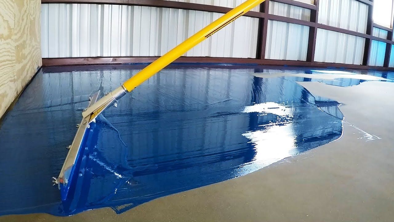 A broom is being used to spread blue paint on a concrete floor.