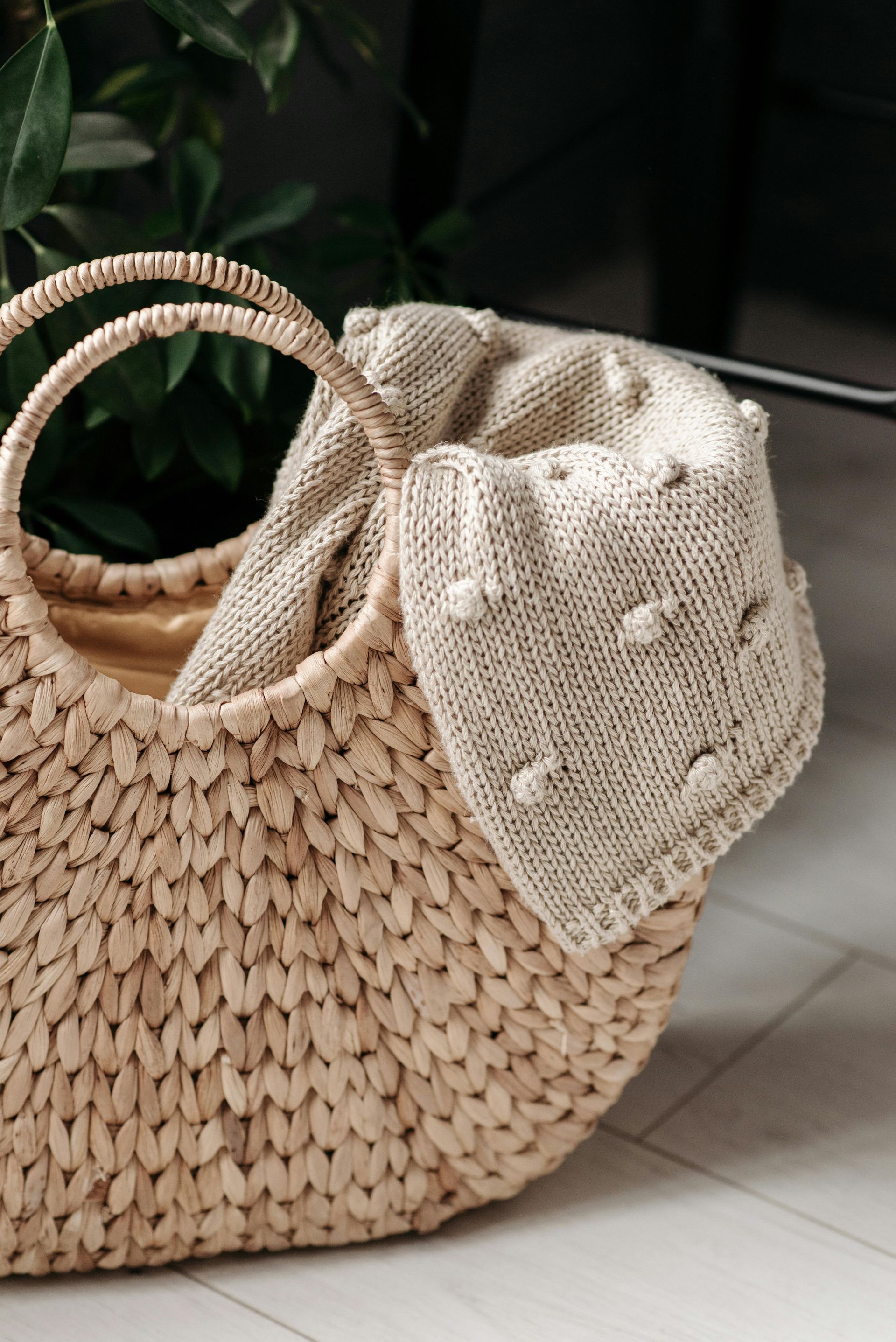 A wicker basket filled with a knitted blanket is sitting on a tiled floor.