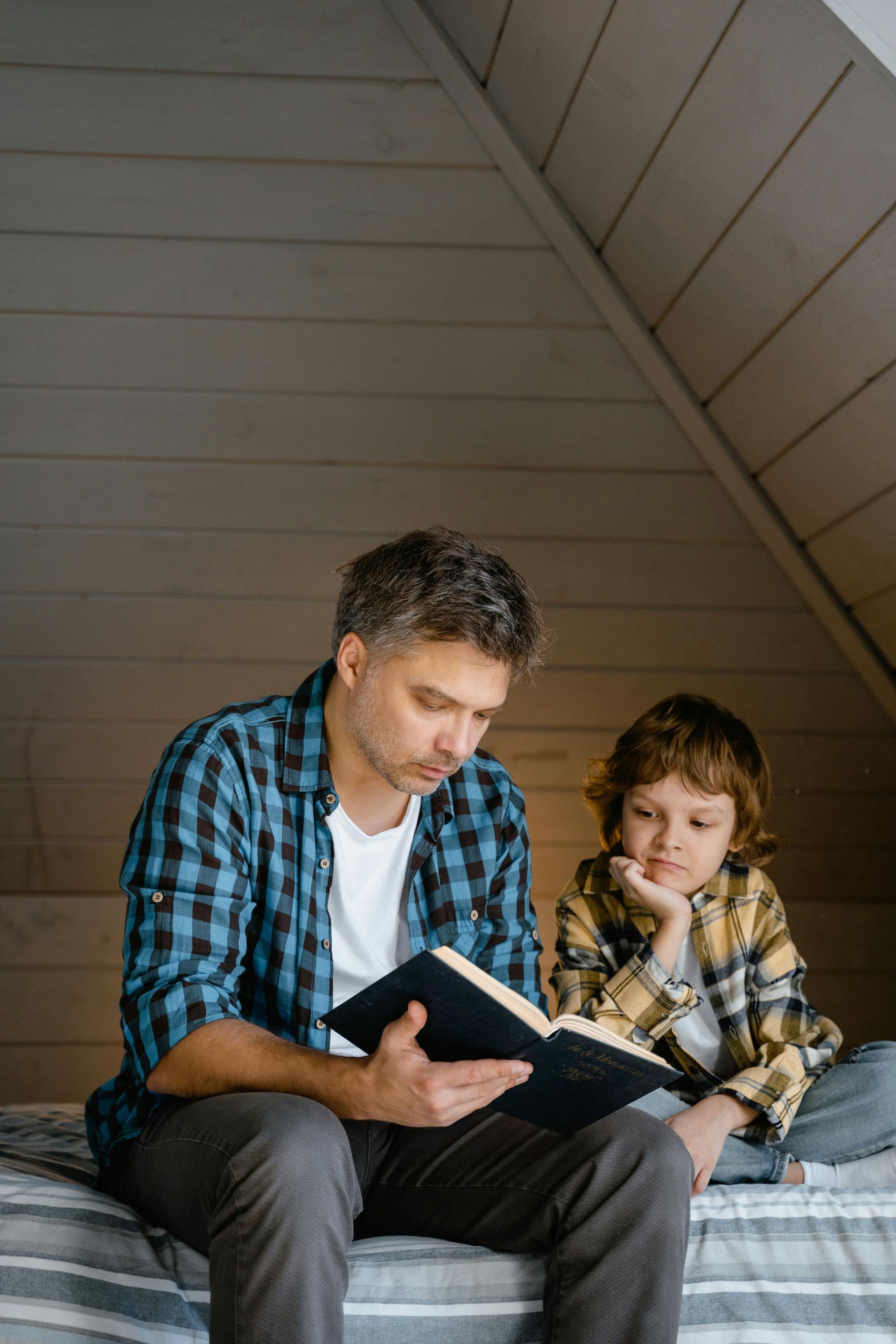 A man and a boy are sitting on a bed reading a book.